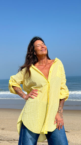 Woman in a yellow shirt standing on a beach with blue sky and ocean.